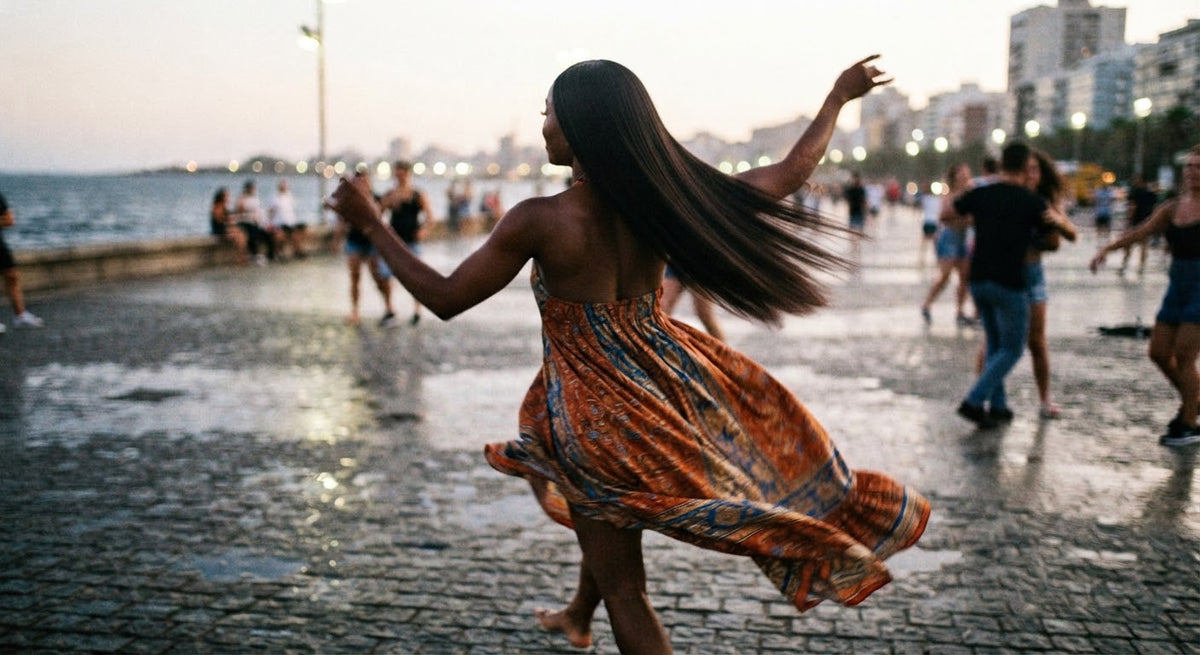 Photographie de dos d'une femme à la peau noire en train de danser sur une promenade pavée en bord de mer, portant une robe longue fluide aux motifs orange et bleus. Ses longs cheveux raides et lisses virevoltent naturellement dans son mouvement sans se soulever ni se décoller à la racine, démontrant une tenue parfaite de la chevelure. L'ambiance est dynamique, capturée sur le vif au crépuscule avec des lumières d'arrière-plan floues, et son visage reste entièrement invisible.